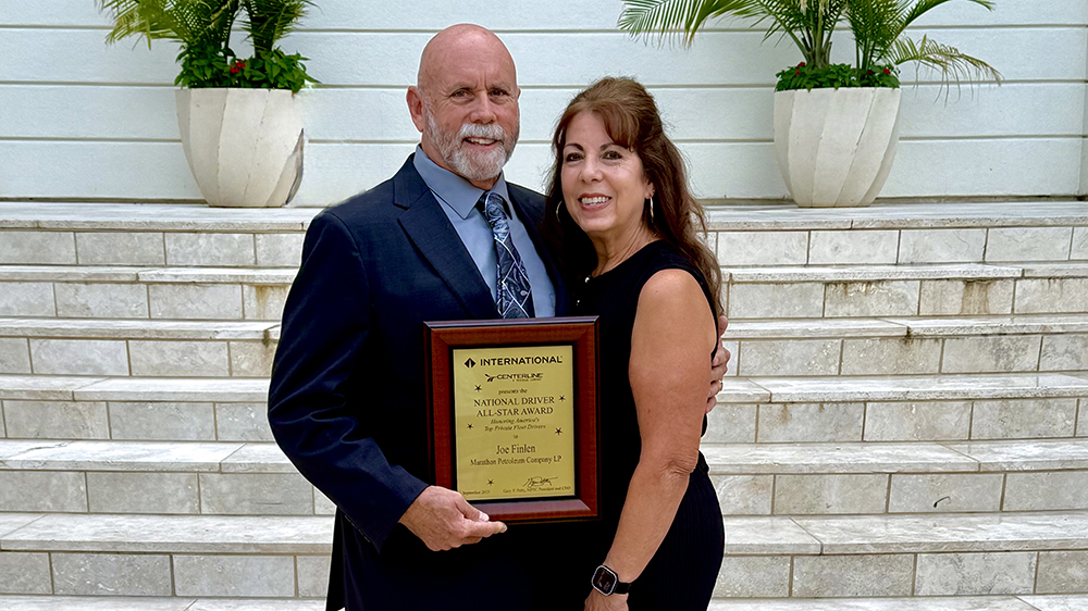 A man and woman posing with the National Driver All-Star Award plaque in front of steps and potted plants.